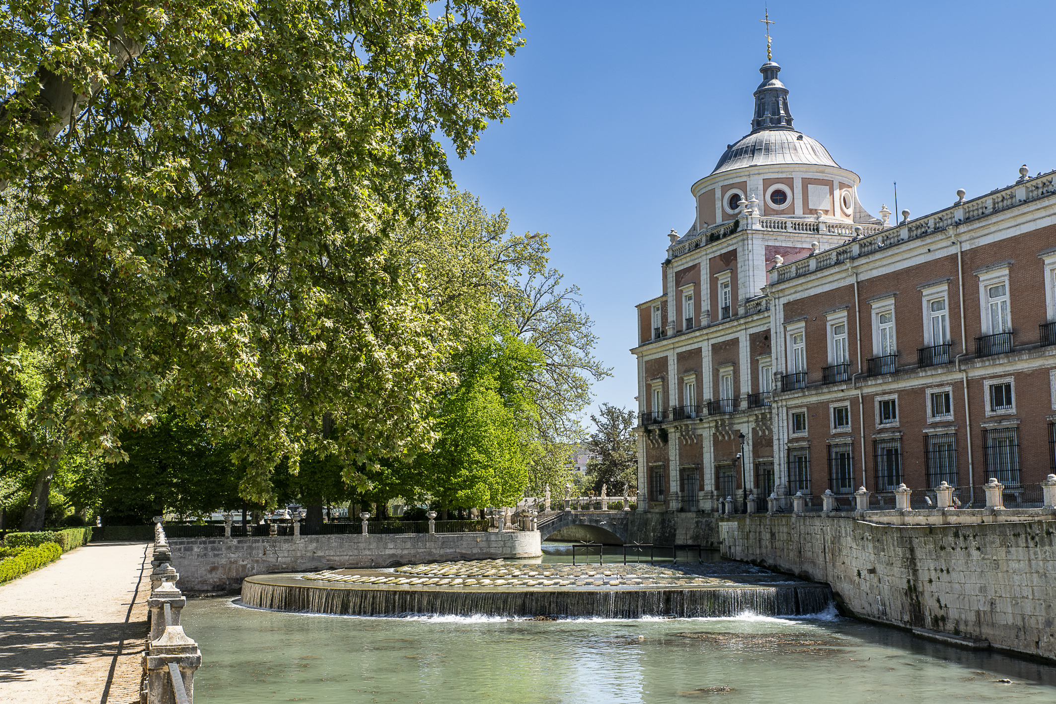 Fotografía actual del palacio de Aranjuez junto a una cascada en el río, entre arboleda.