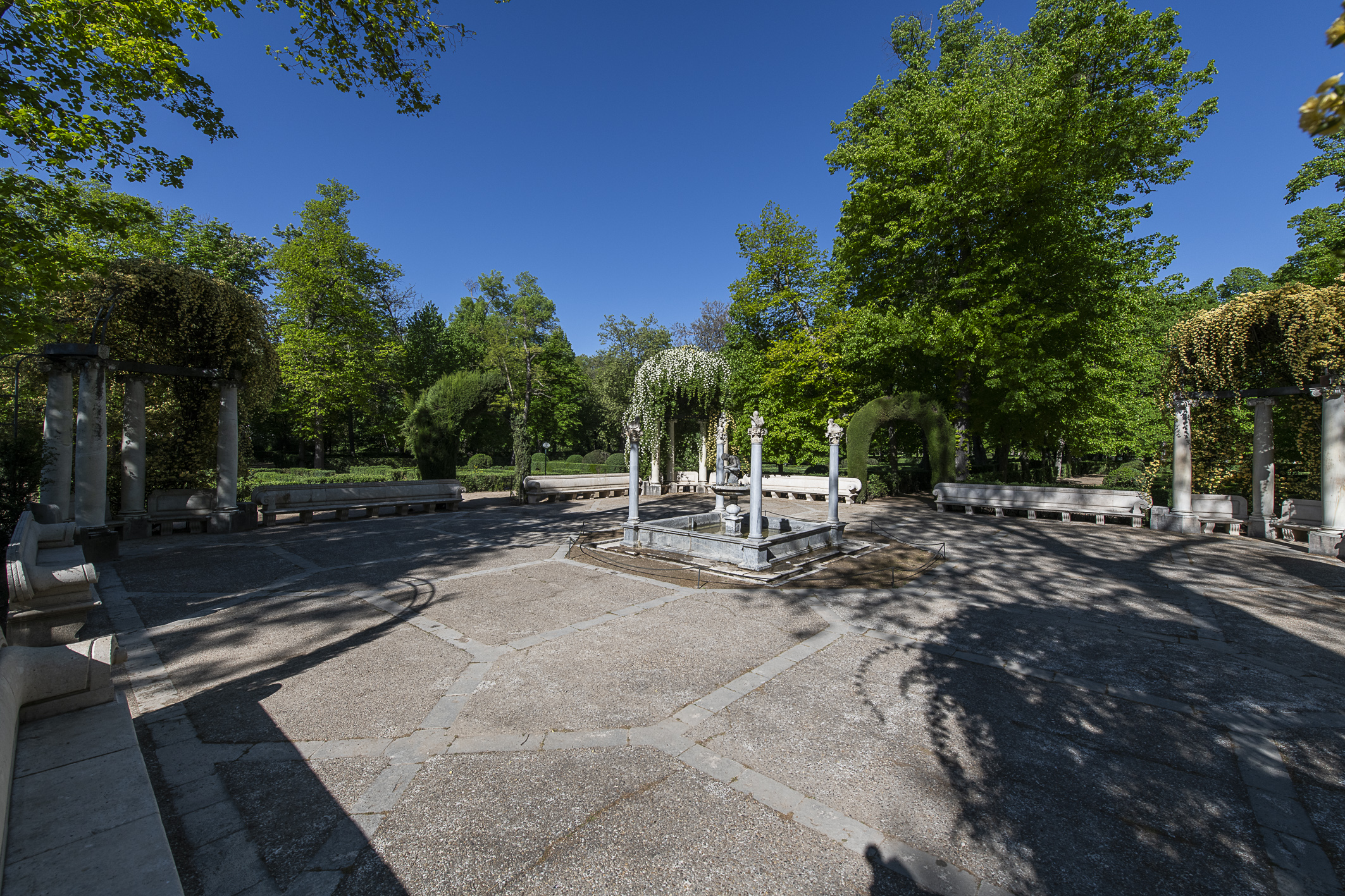 Fotografía de una glorieta empedrada con columnas, pérgola y una pequeña fuente central.
