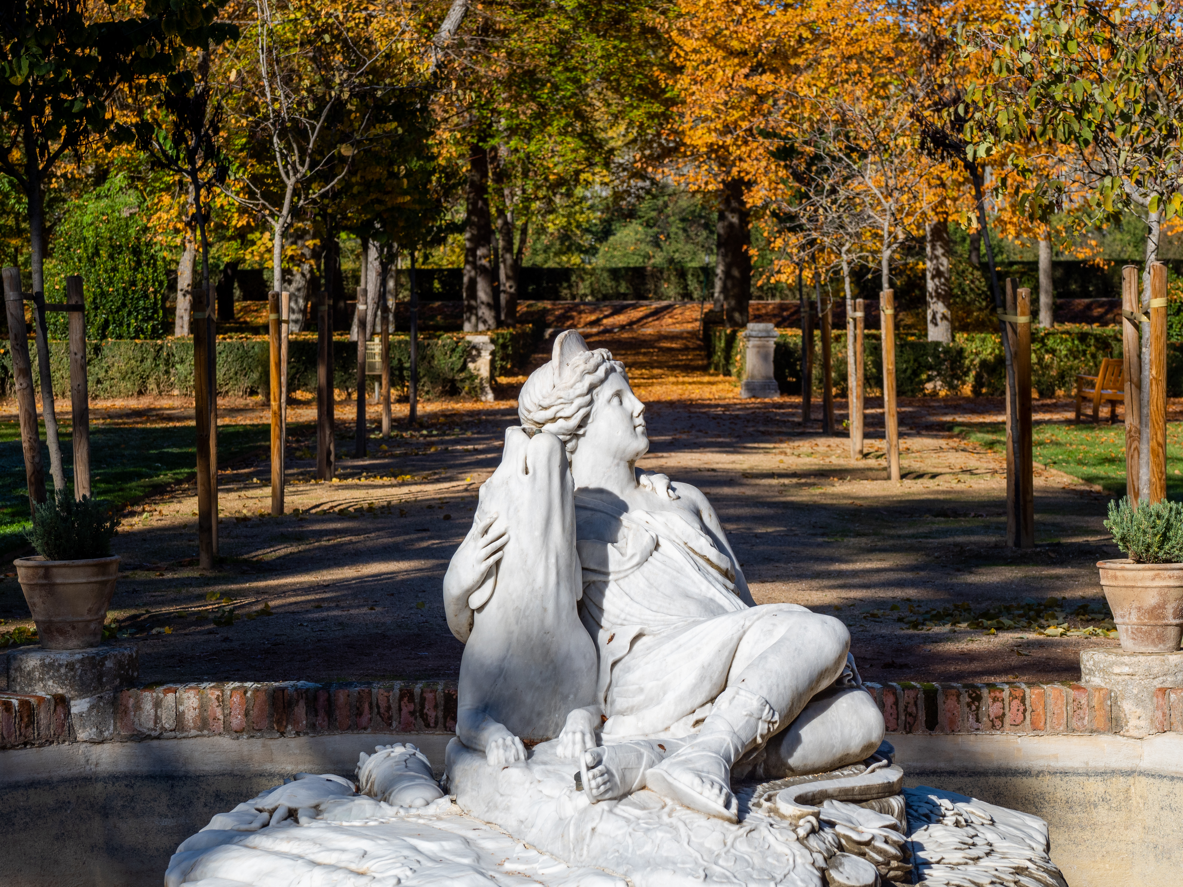 Detalle de una escultura de mármol con figuras recostadas en el jardín, con arbolado otoñal.