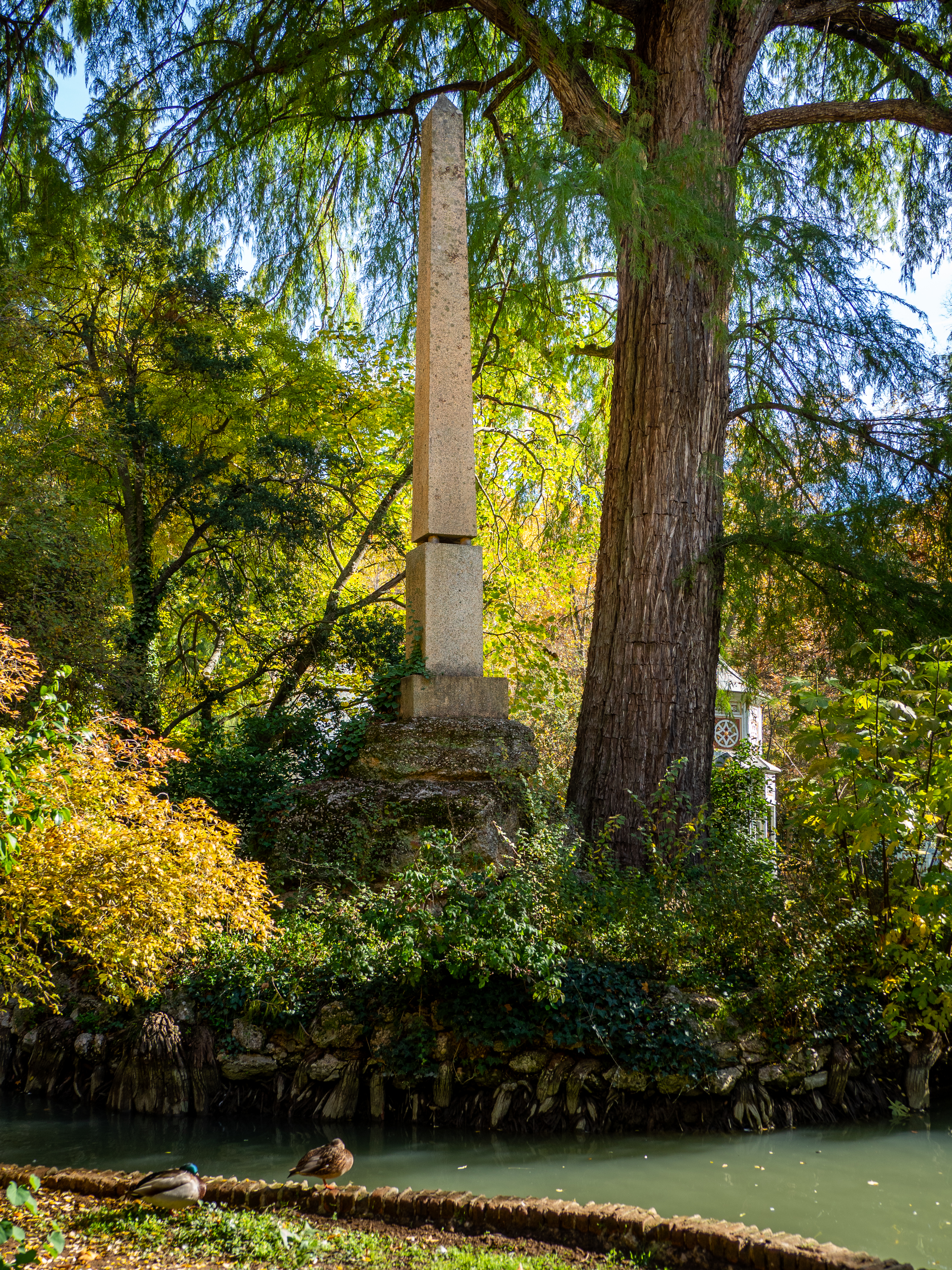 Fotografía actual de un obelisco junto al estanque, bajo un gran árbol otoñal.