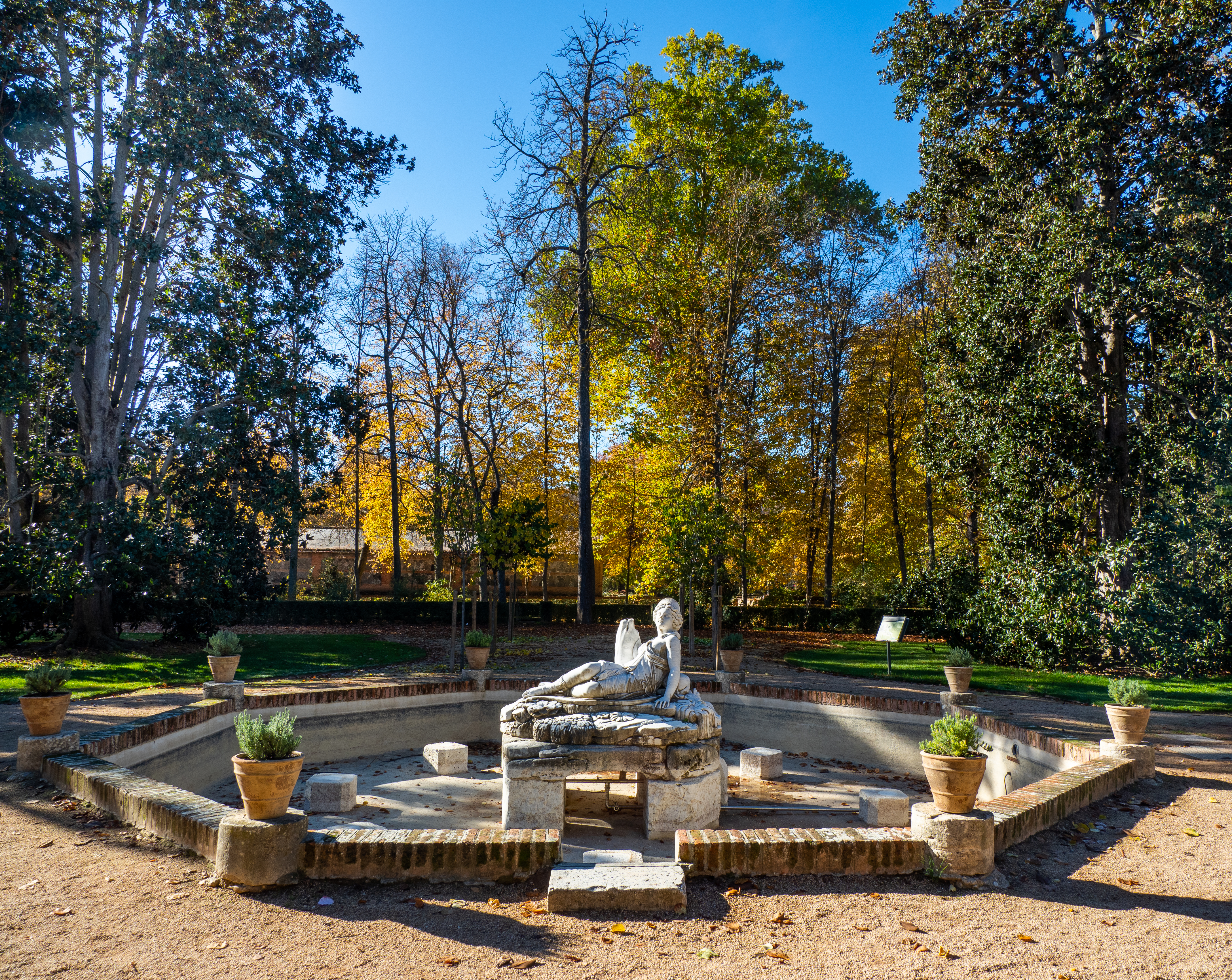 Escultura yacente de mármol en una fuente hundida del jardín, rodeada de arbolado otoñal.