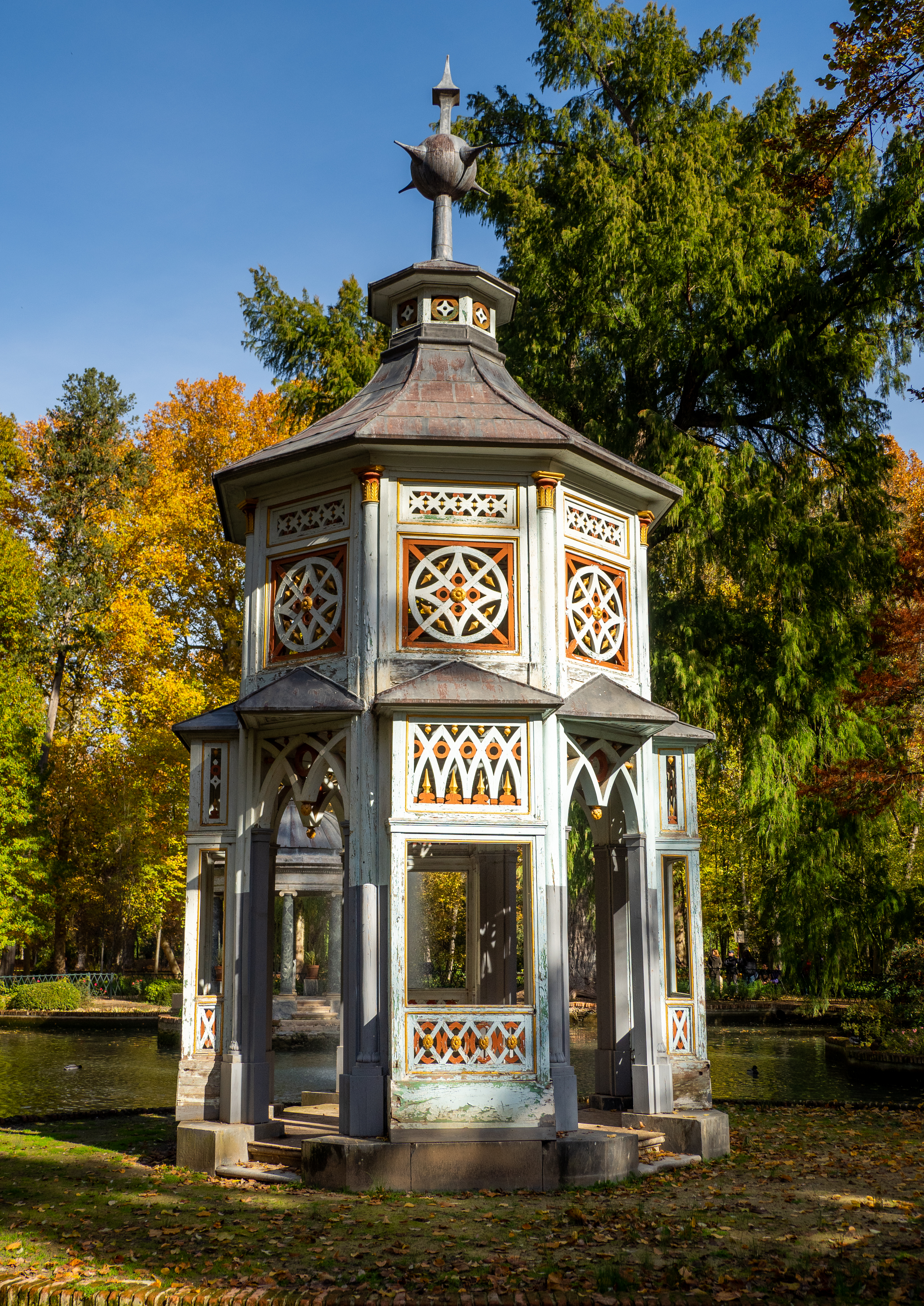 Fotografía de un templete chinesco blanco junto a un estanque, con arbolado otoñal.