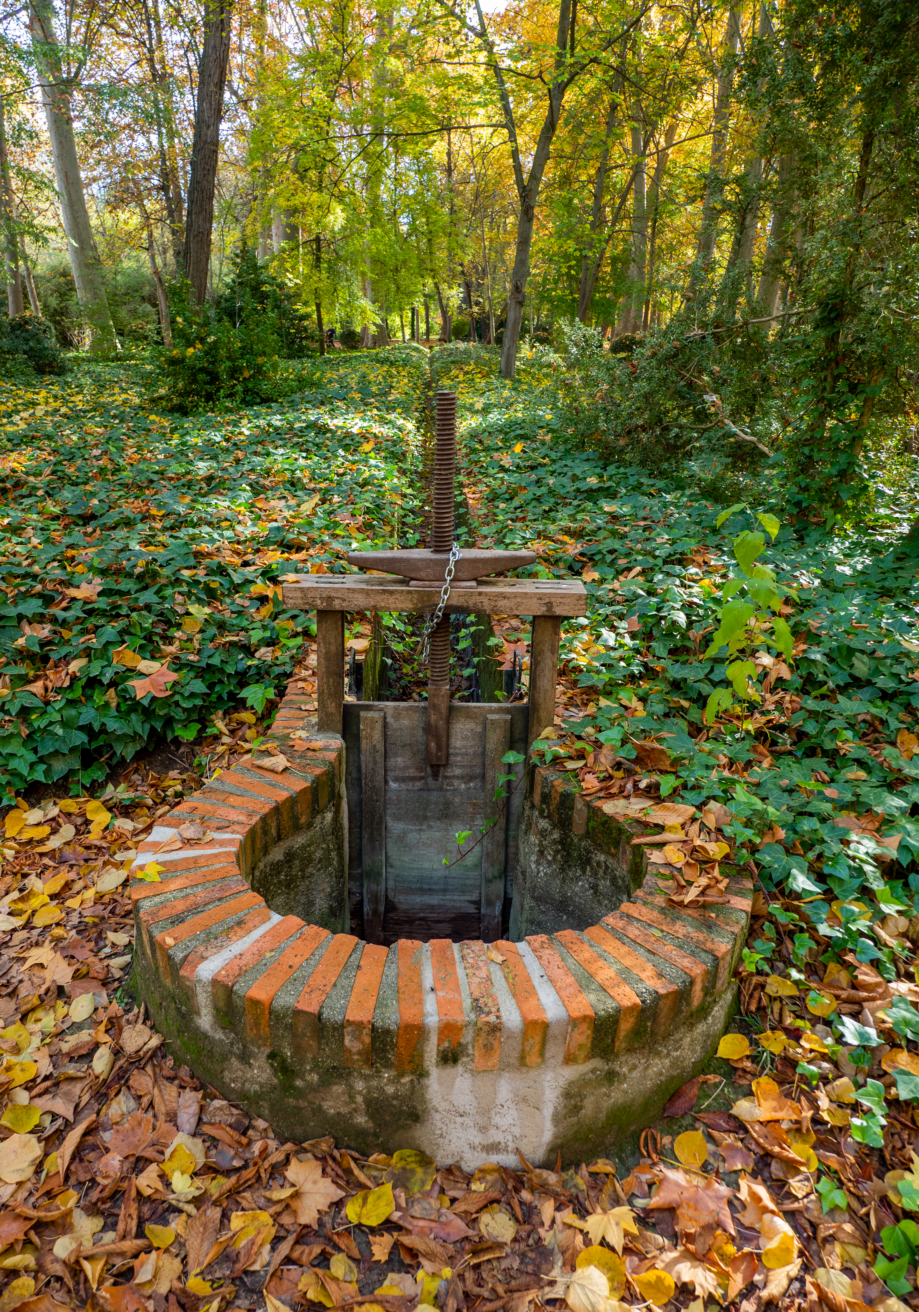 Fotografía de una compuerta circular de ladrillo con husillo de hierro en el bosque.
