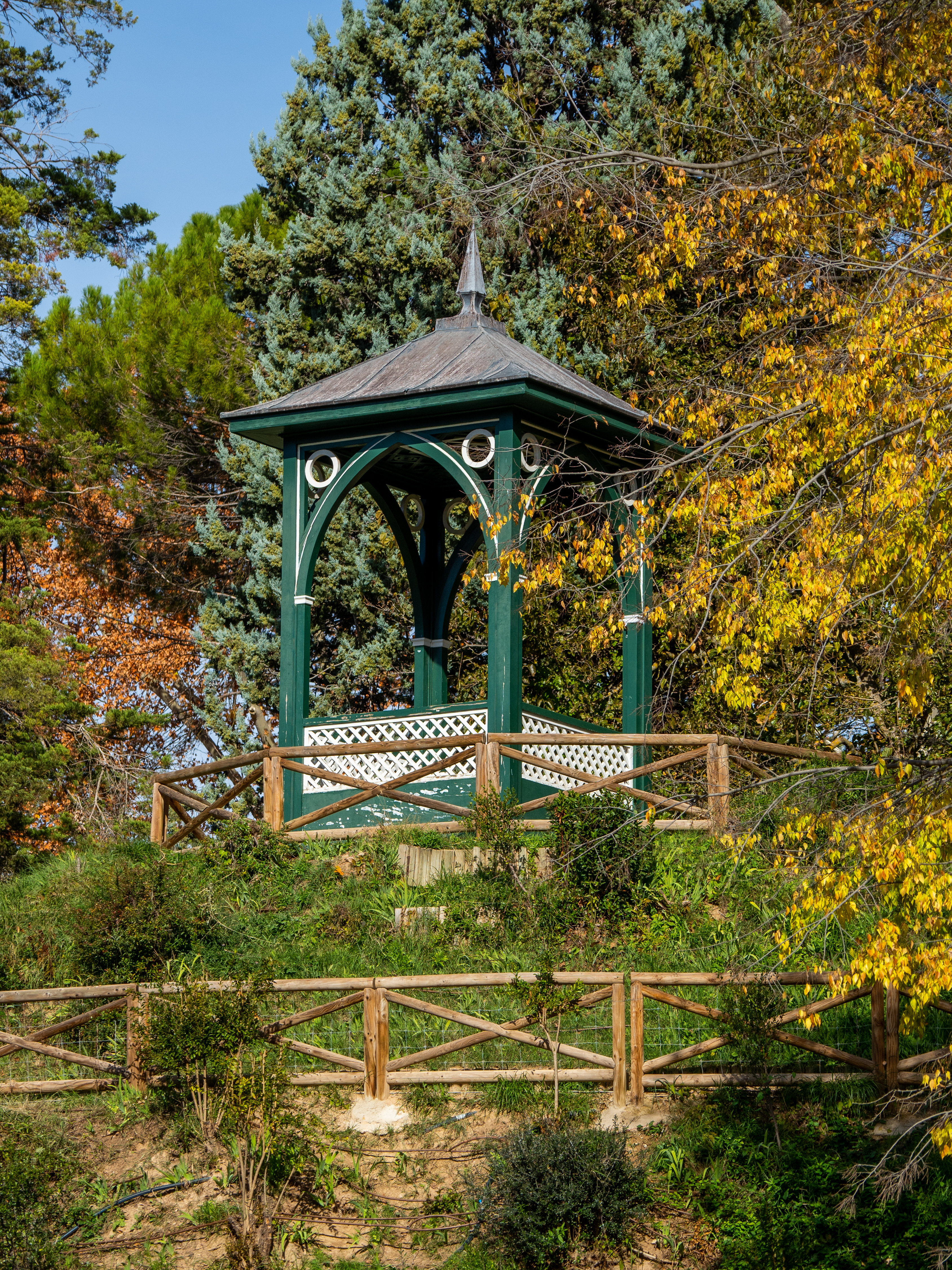 Fotografía de un templete hexagonal verde con balaustrada calada, entre vegetación otoñal.